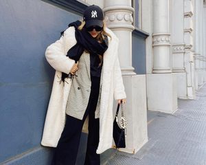 Audrey Kate Lopez com casaco peludo branco, blazer xadrez, cachecol, chapéu do New York Yankees, calça e bolsa preta