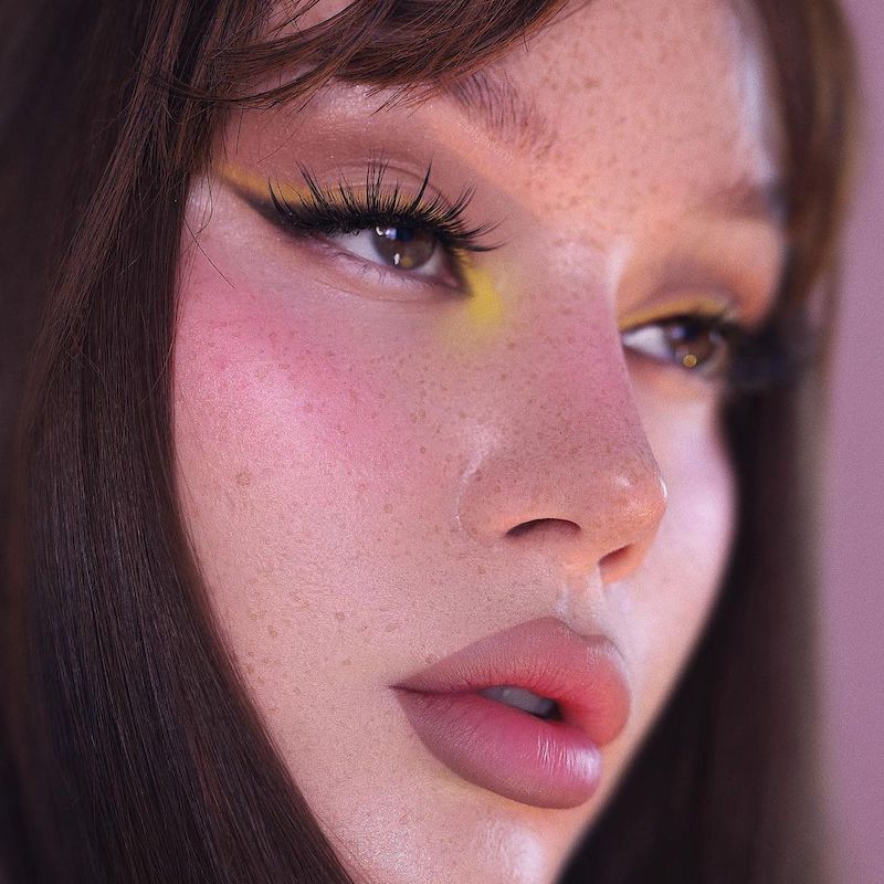Modelo em maquiagem com pálpebras amarelas, cílios pintados e lábios para baixo