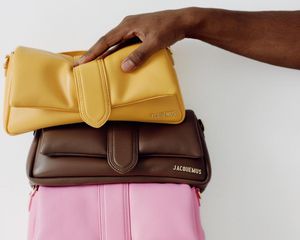 Mão tocando uma pilha de bolsas Jacquemus coloridas amarelas, marrons e rosa