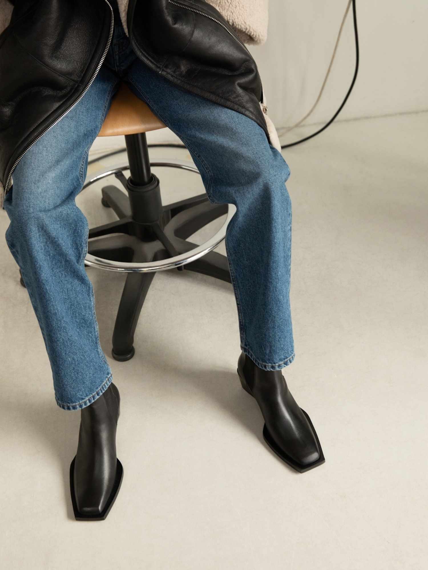 Uma pessoa próxima de uma pessoa em jeans com pernas retas, uma jaqueta de couro feita de couro cortado e botas pretas Chelsea com um dedo do pé quadrado