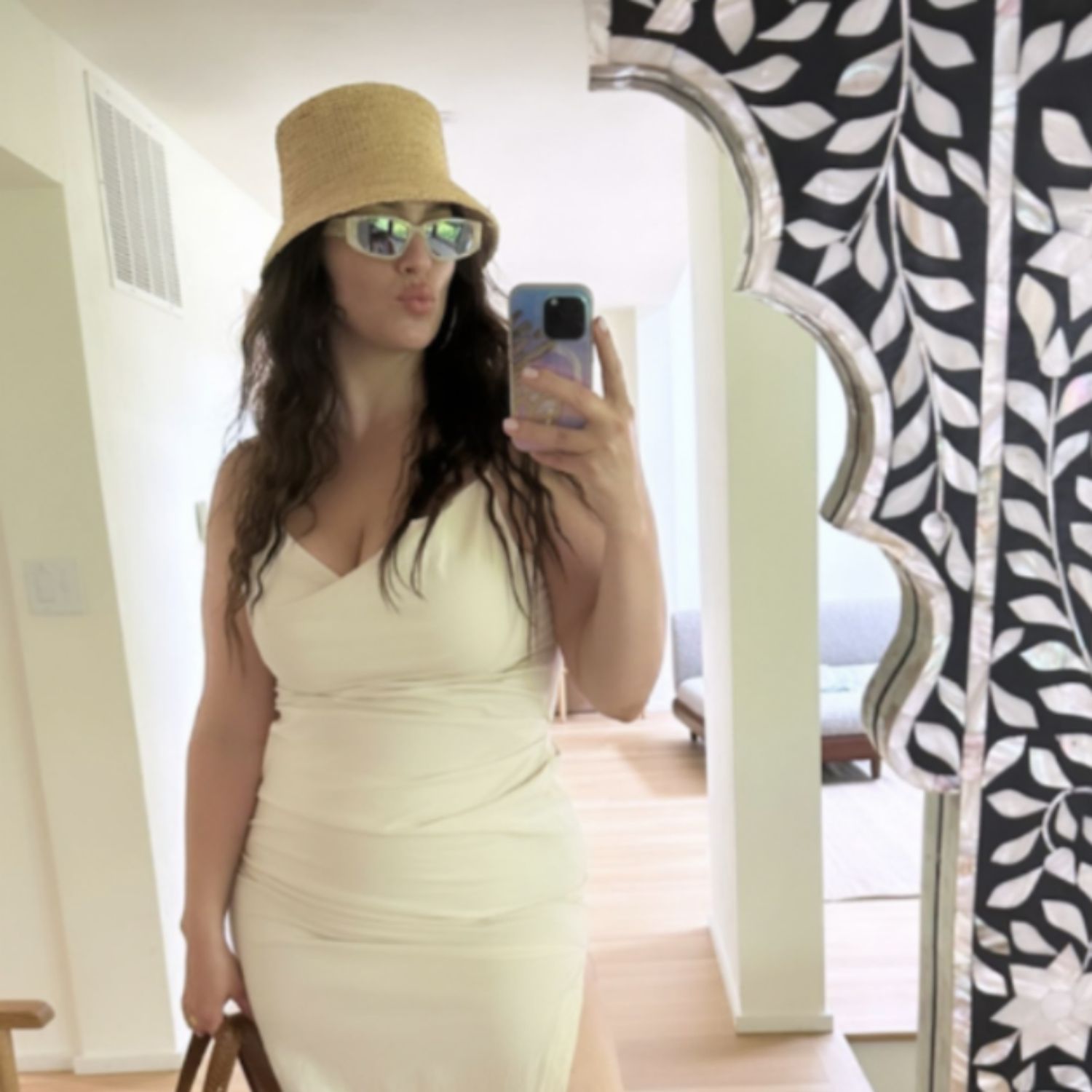 Ashley Graham faz uma selfie em um espelho em um vestido branco, óculos escuros e um ódio de palha