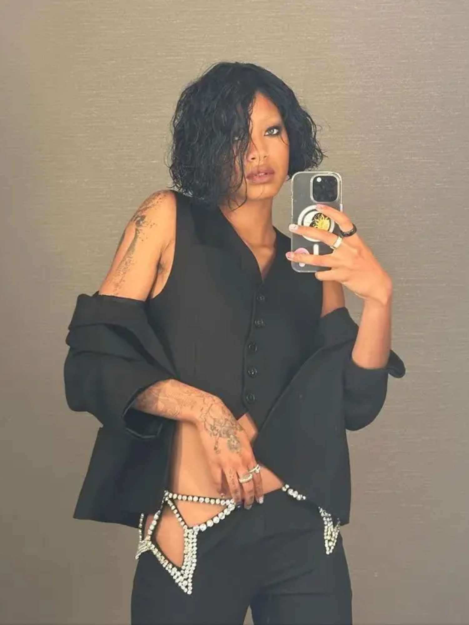 Willow Smith tira uma selfie no espelho com um colete preto de botão, blazer de braço, calça recortada de strass prateado, anéis e maquiagem esfumada nos olhos
