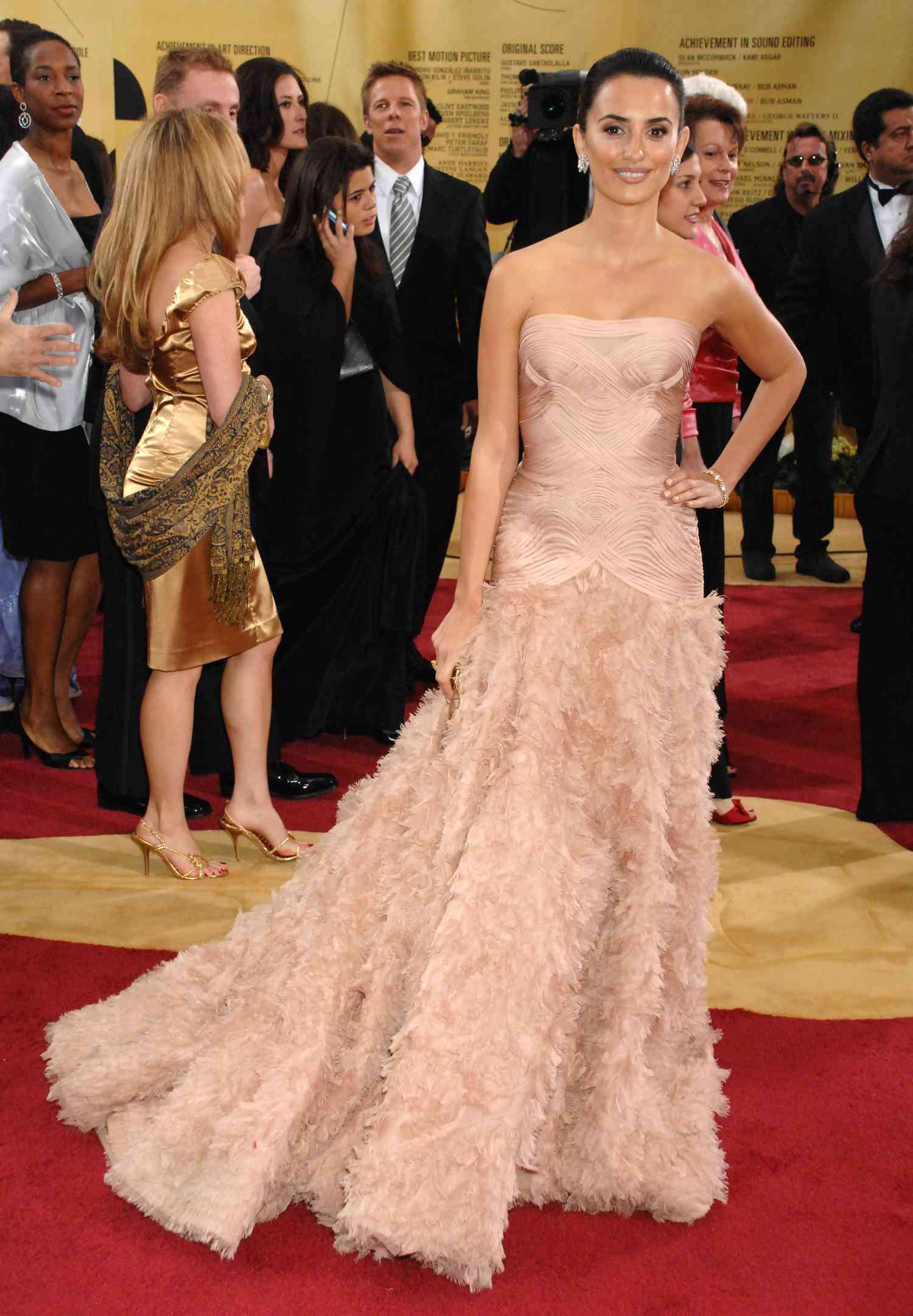 Penelope Cruz fica na rua no cenário de muitas pessoas. Ela está vestida com um vestido rosado sem mangas com uma saia cortada. O cabelo é colocado em um pacote apertado, uma mão está na coxa.