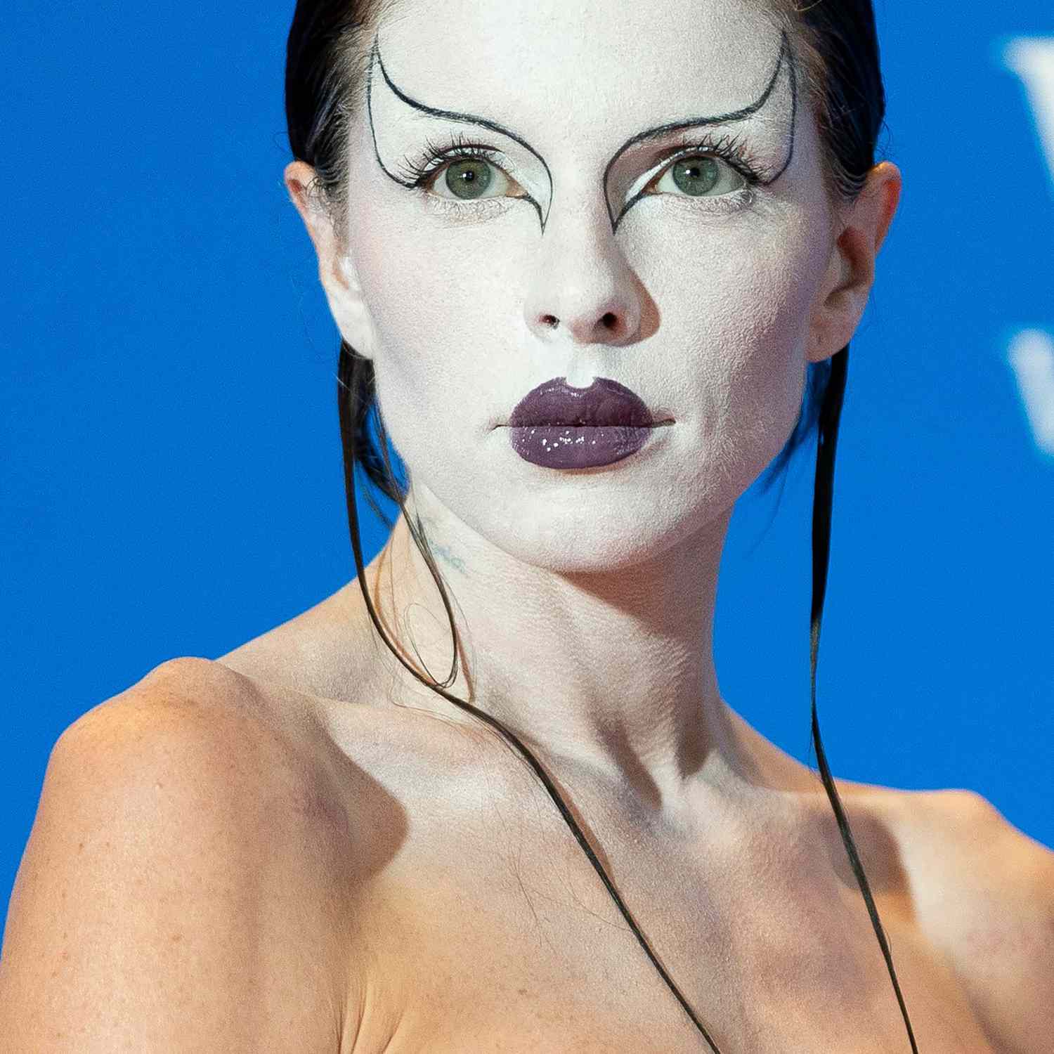 Julia Fox em maquiagem branca com asas pretas finas, descrevendo os dois olhos e o batom roxo