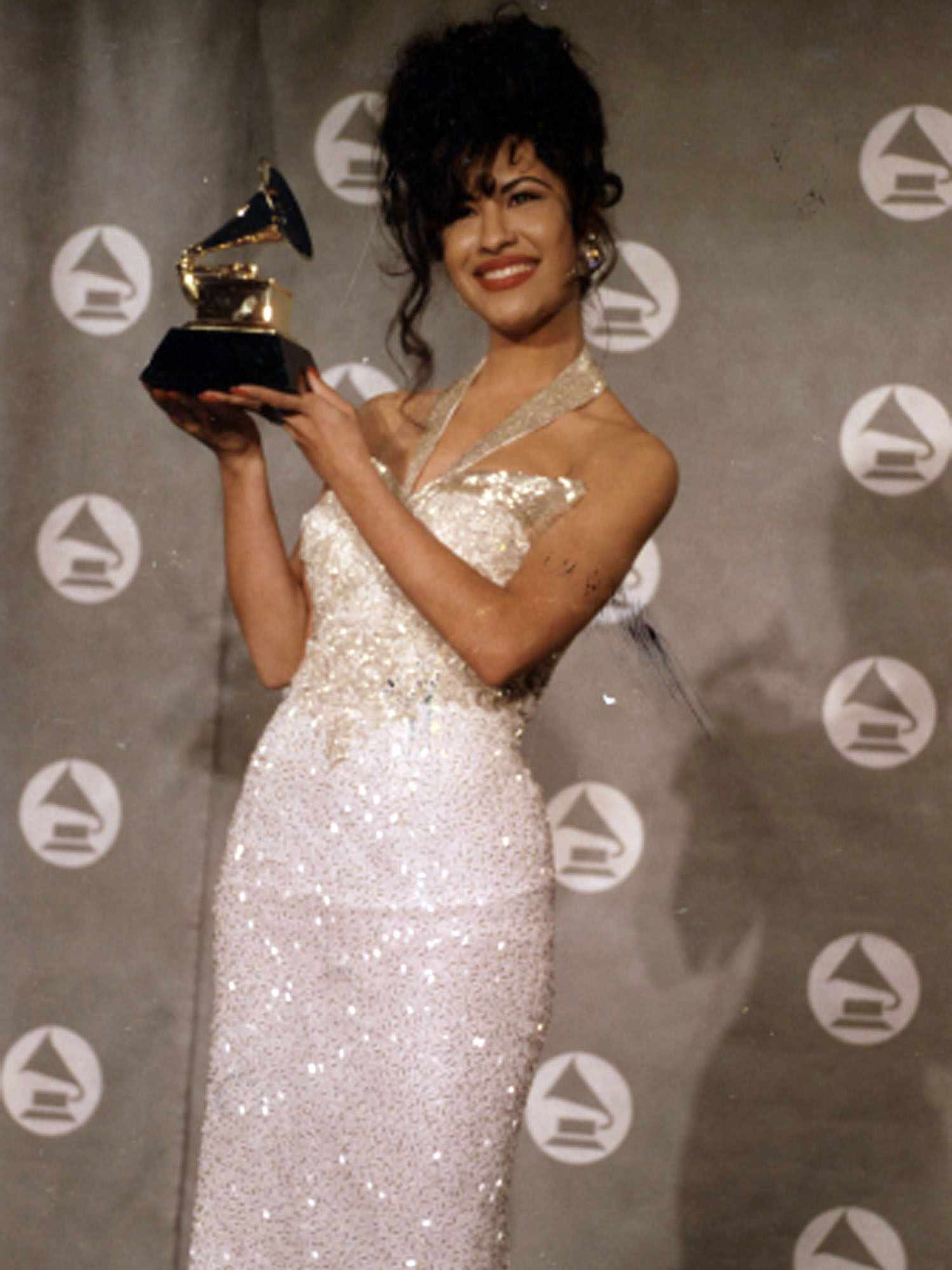 Selena em um vestido prateado com brilhos segura em suas mãos seu prêmio Grammy de 1994.