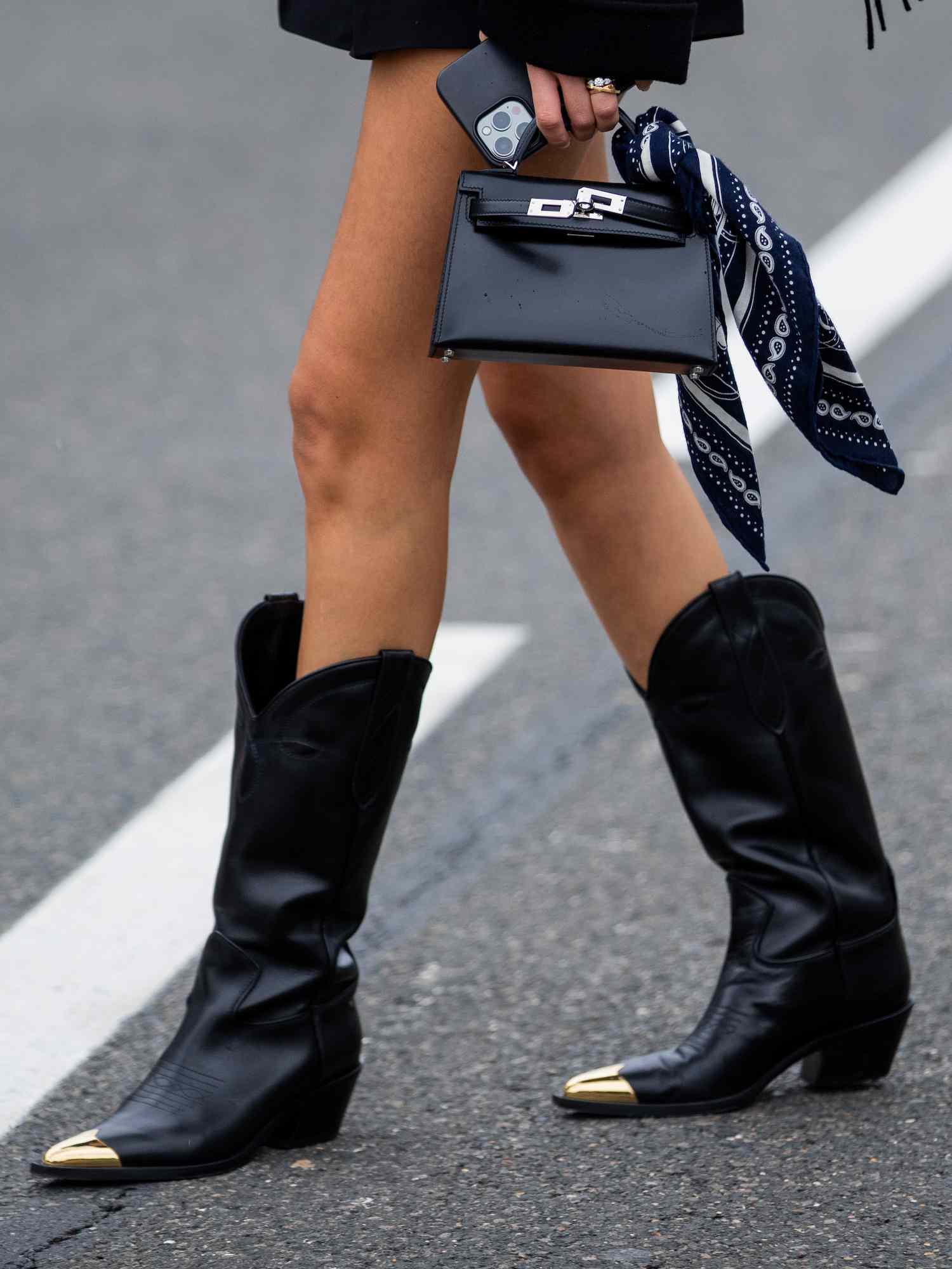 Uma mulher próxima em uma jaqueta com uma franja, uma bolsa preta de Hermes e botas de couro preto no estilo de Western com uma malha de ouro < man> botas pretas ava da linha