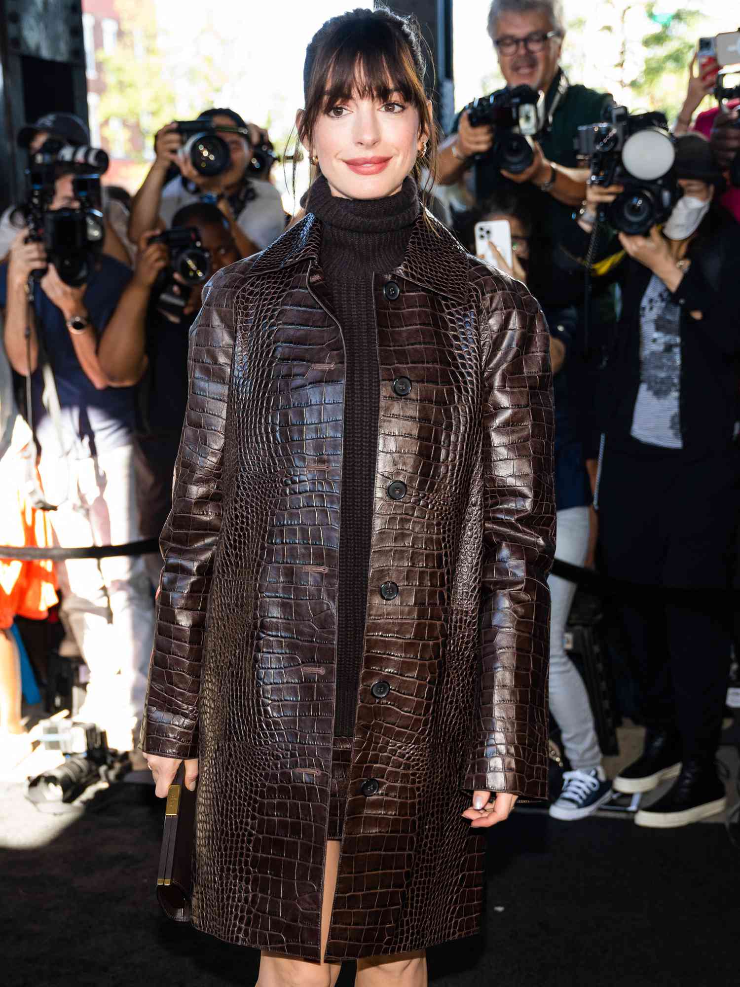 Anne Hathaway com casaco marrom com efeito crocodilo da Michael Kors, gola alta marrom chocolate e penteado com franja ondulada
