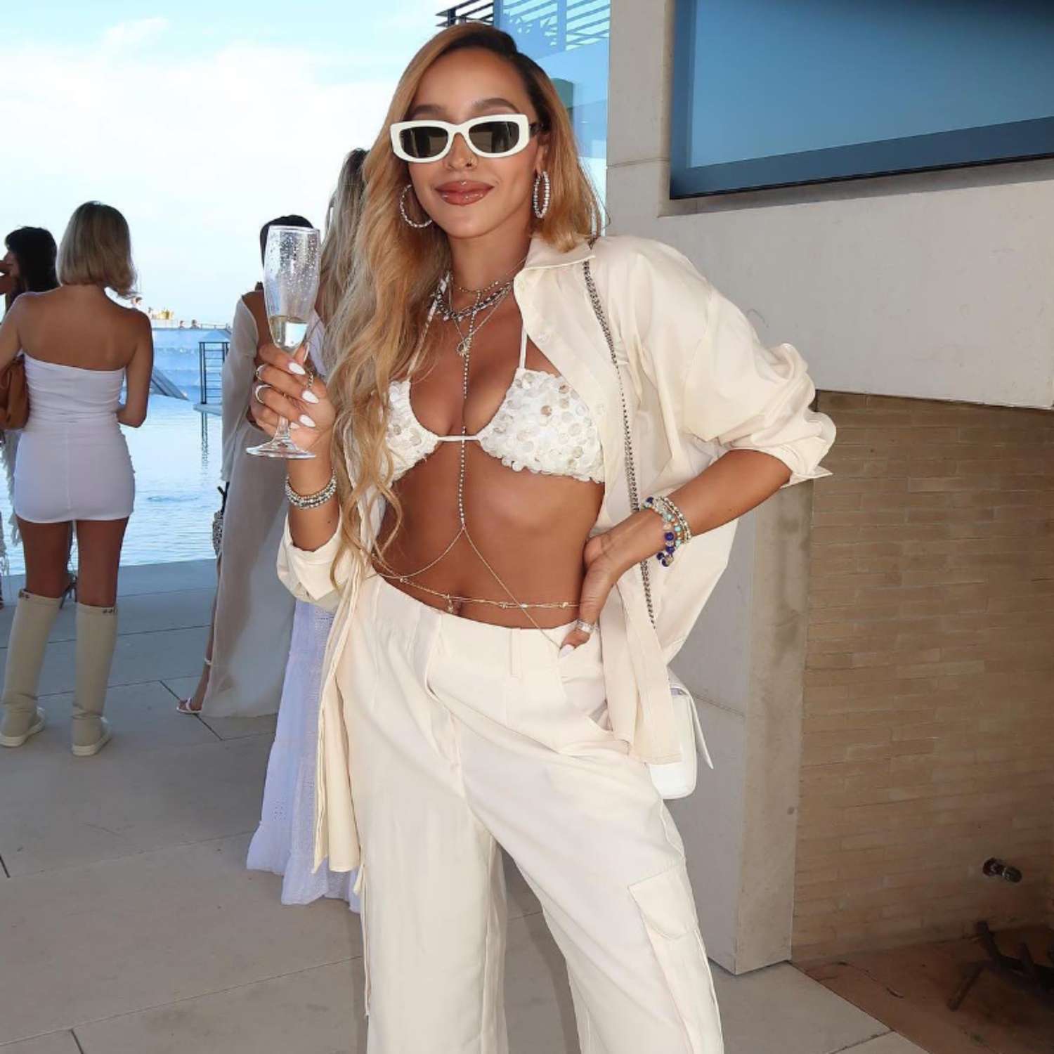 Tinashe posa com uma taça de champanhe em uma festa usando óculos escuros brancos, body, blusa de banho branca, calça branca e camisa de botão branca.