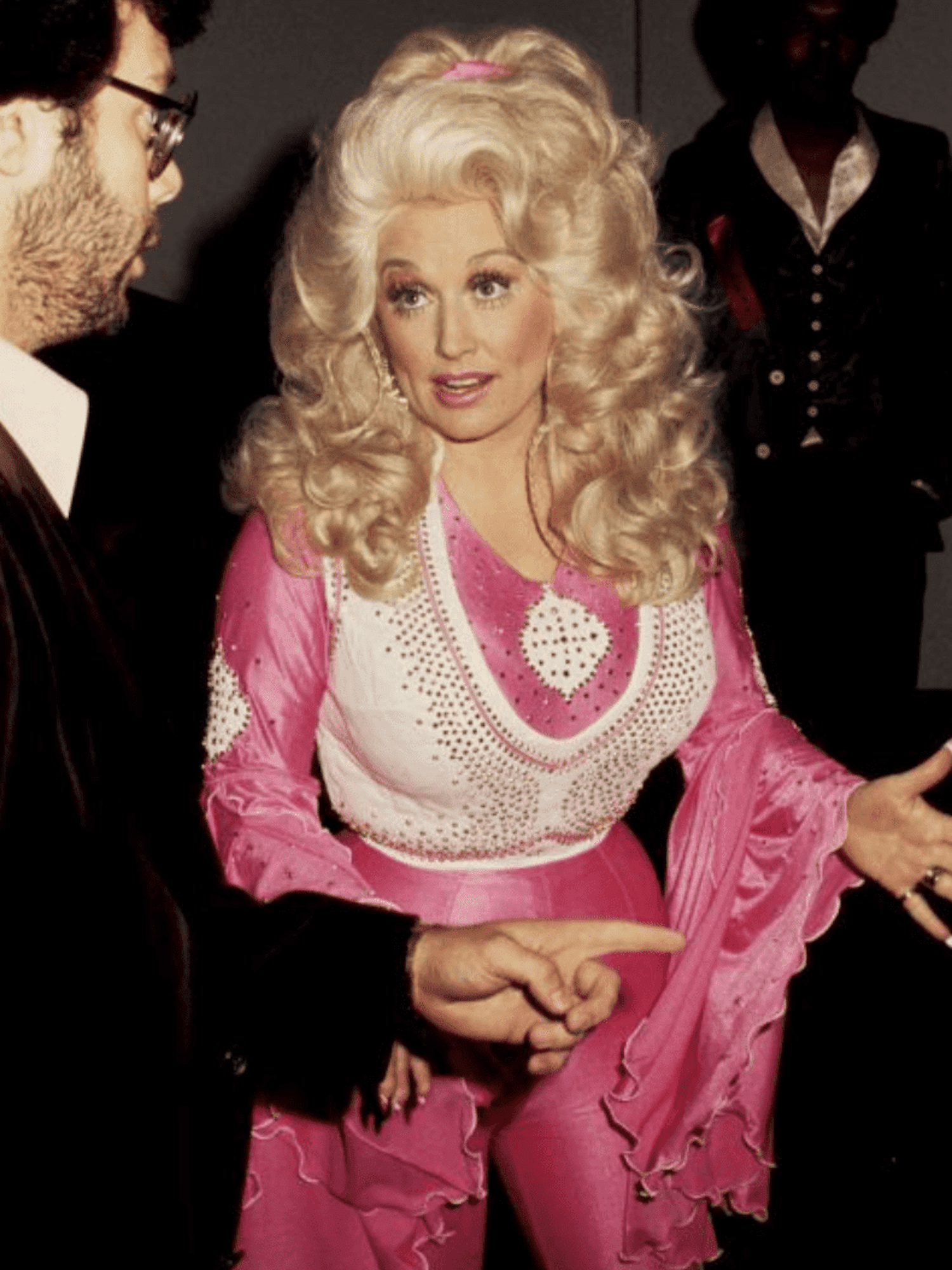 Dolly Parton em macacão rosa e branco no estilo de cowboy e com cabelos encaracolados volumosos