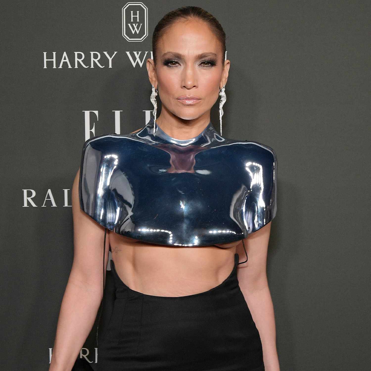 Jennifer Lopez em um babador de metal prateado, brincos de prata pendurados e unhas cromadas em ouro champanhe